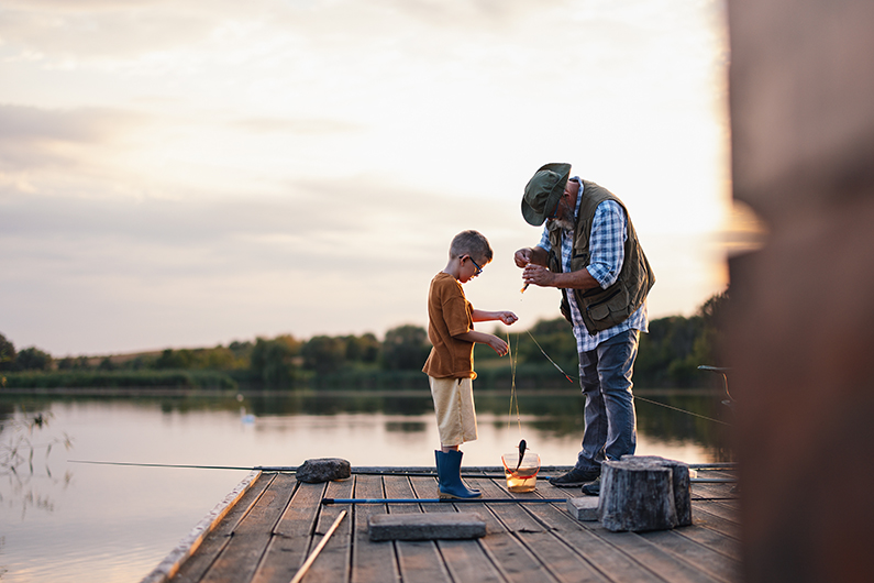 A man and a child standing on a wooden dock by a calm lake at sunset, working together with fishing lines and a bucket between them.