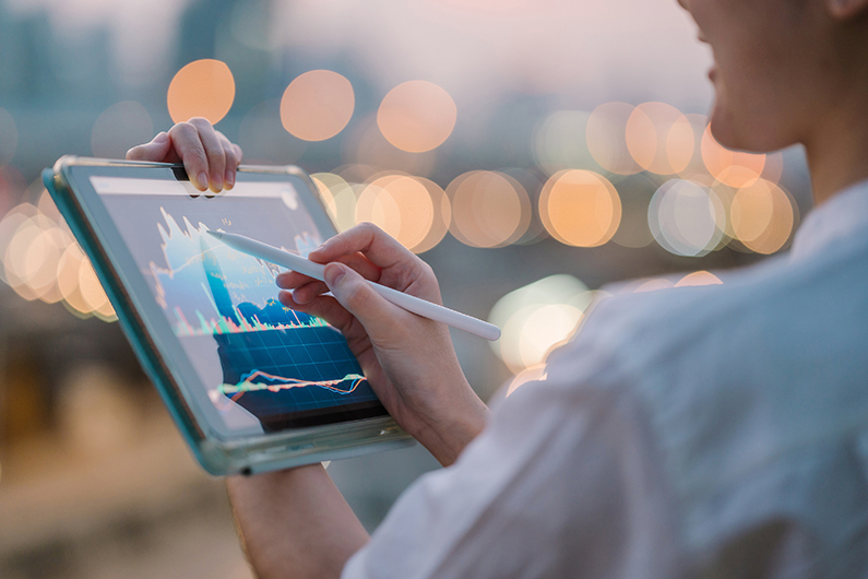 Person using a stylus to interact with a tablet displaying various financial graphs and charts against a blurred cityscape background with bokeh lights.