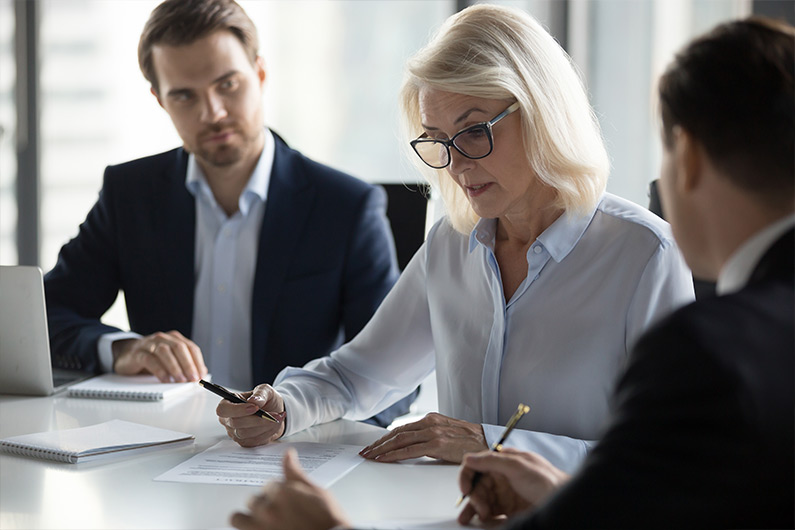 meeting between two guys in suits and an older women with blond hair and glasses reading over paperwork holding a pen